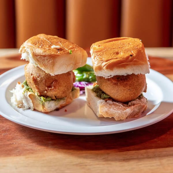 Two vada pavs, Indian potato fritter sliders, with green chutney and red cabbage on a white plate.