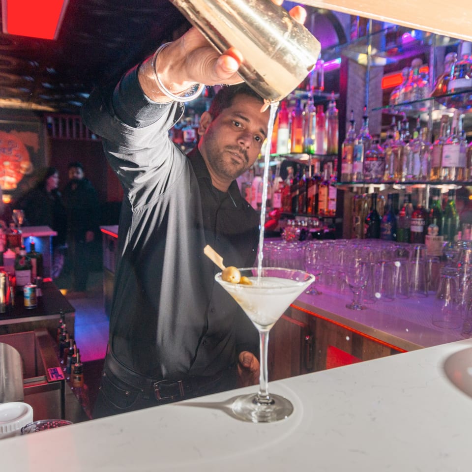 Bartender pouring a martini from a shaker into a glass with olives at Taste of Mumbai.