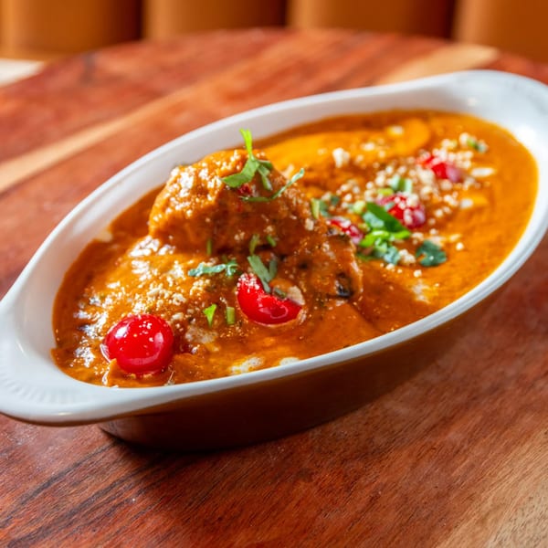 A white oval dish filled with a rich orange Indian curry, garnished with fresh herbs and red berries.