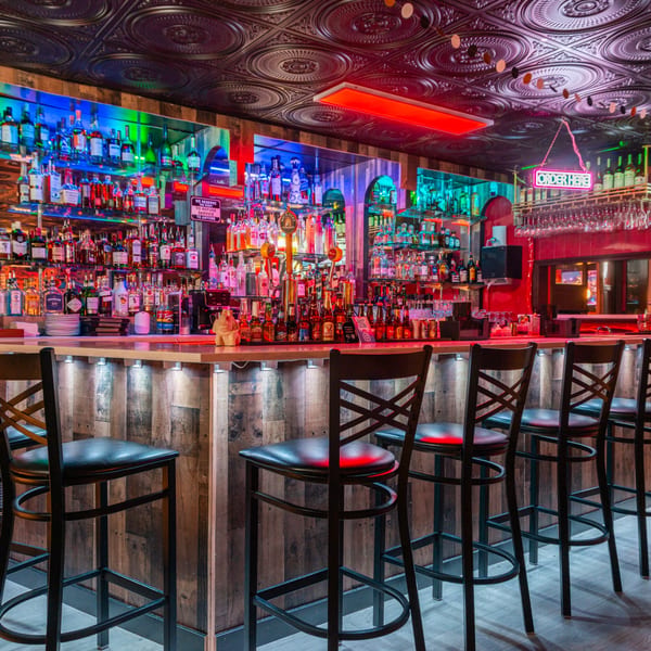 A vibrant bar with colorful lighting, rows of liquor bottles, and empty bar stools at Taste of Mumbai.