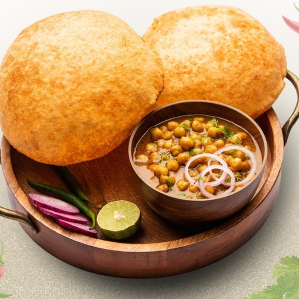 Two golden-brown puris, a bowl of chole, sliced red onions, green chilies, and lime on a wooden tray.
