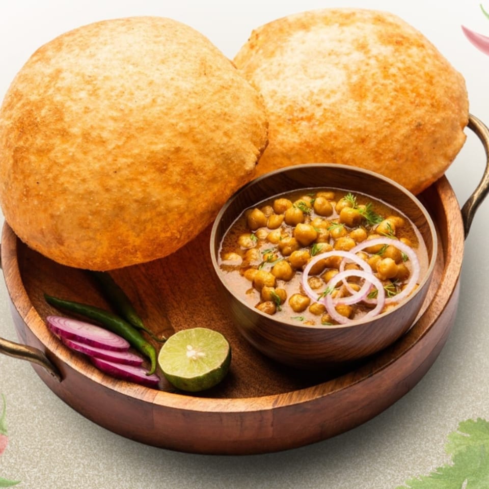 Two golden-brown puris, a bowl of chole, sliced red onions, green chilies, and lime on a wooden tray.